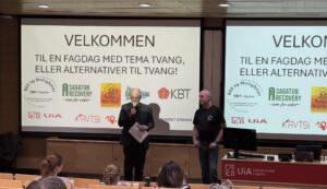 Bilde av to menn som snakker foran en forsamling. I bakgrunnen er det et lerret hvor det st¨r "Velkommen til en fagdag med tema tvang eller alternativer til tvang"